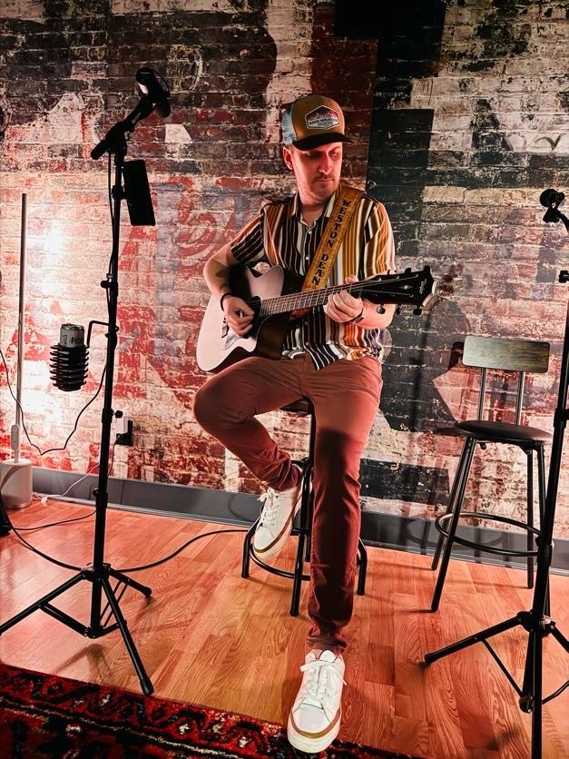 Weston Dean performing on acoustic guitar against a painted brick wall